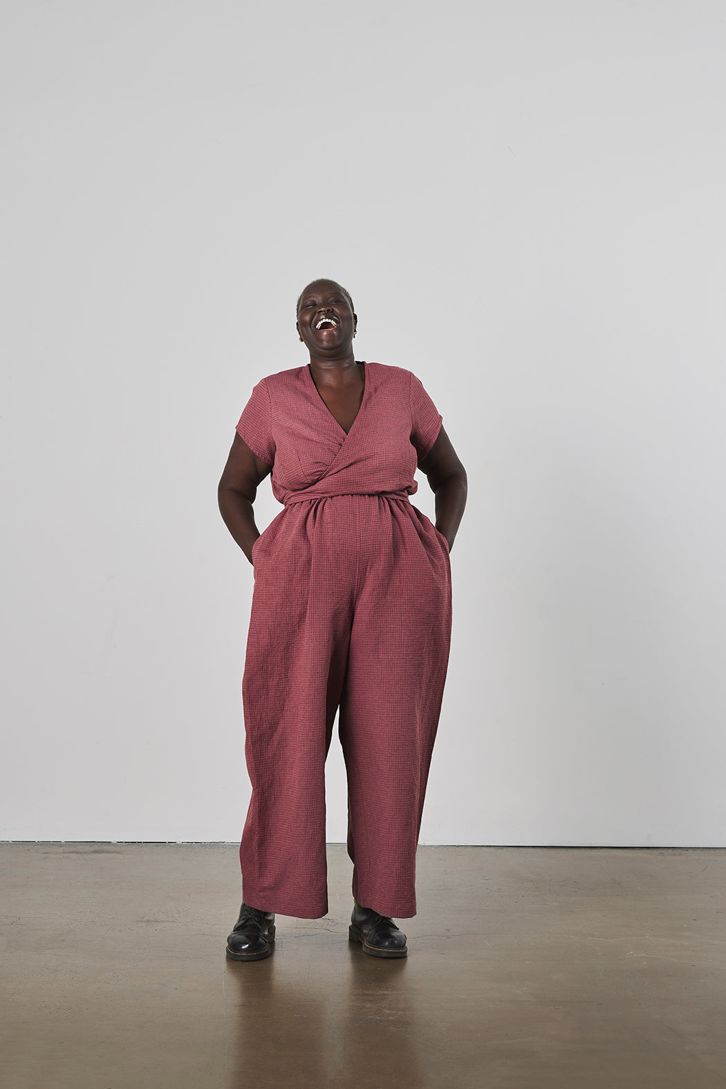 Laughing person wearing a maroon jumpsuit with hands in pockets standing against a white wall.
