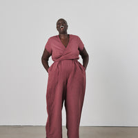 Laughing person wearing a maroon jumpsuit with hands in pockets standing against a white wall.